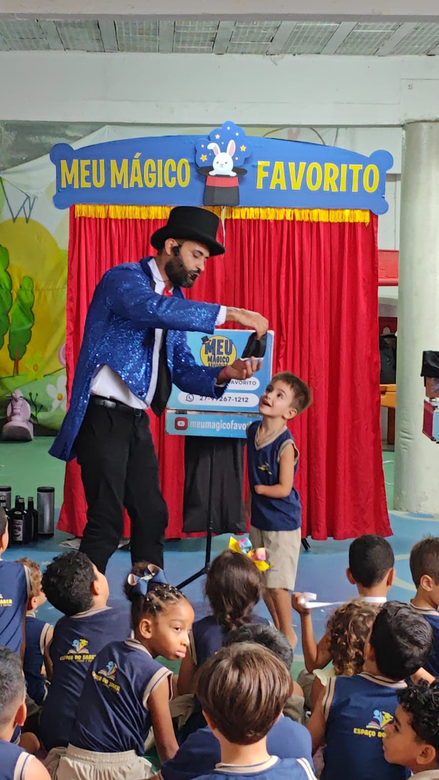 Criança sorrindo ao lado do mágico durante um truque no palco da escola