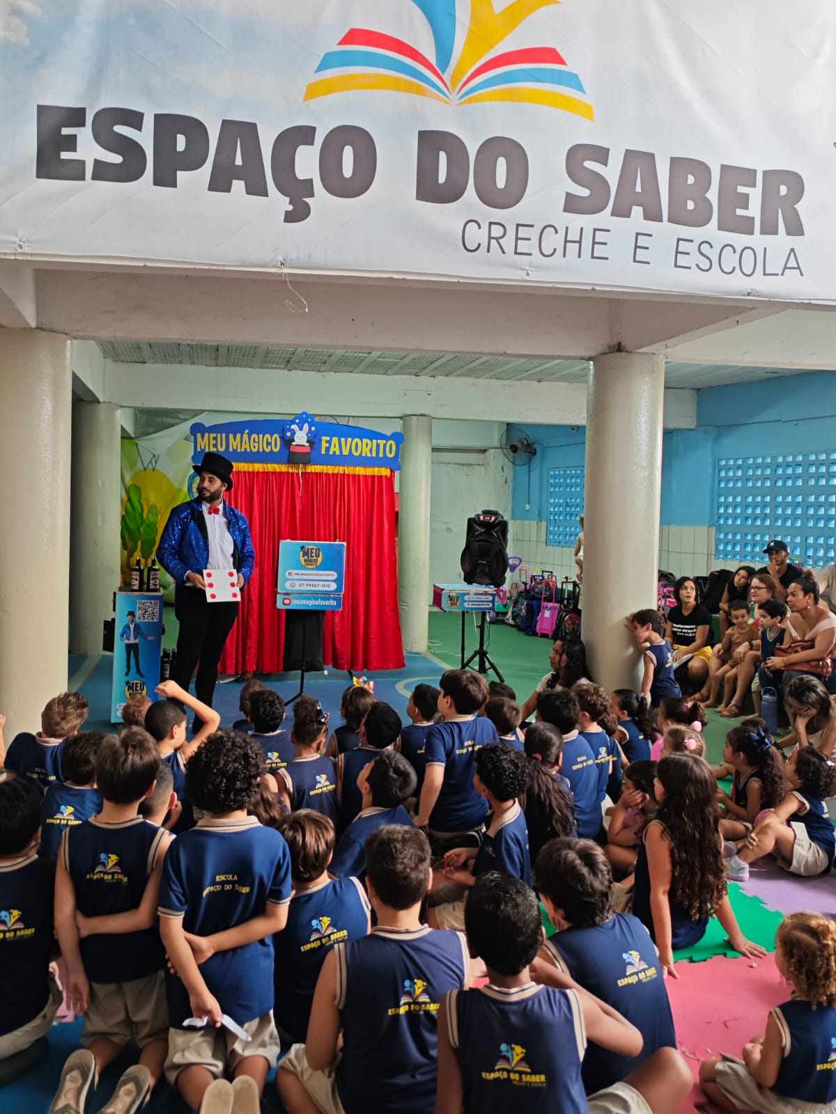 Mágico se apresentando no palco da escola Espaço do Saber, com cortina vermelha e faixa da marca Meu Mágico Favorito