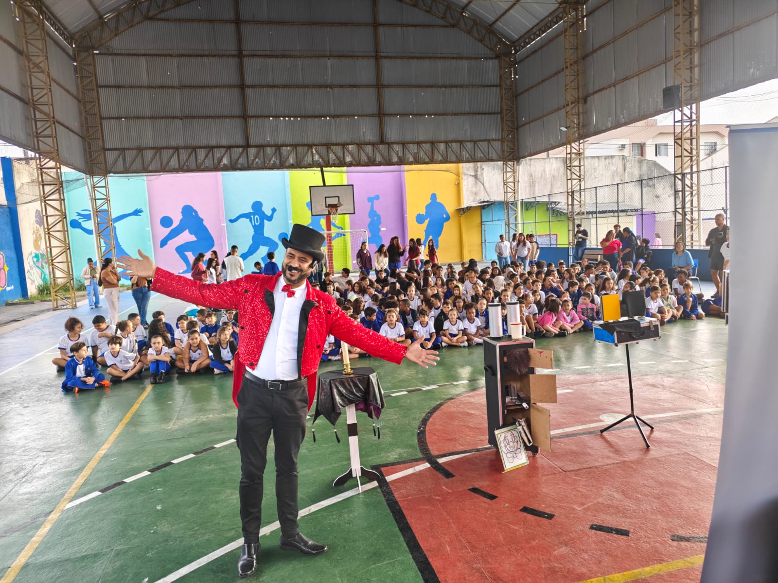 Mágico de paletó vermelho de braços abertos diante de uma quadra escolar lotada de crianças encantadas